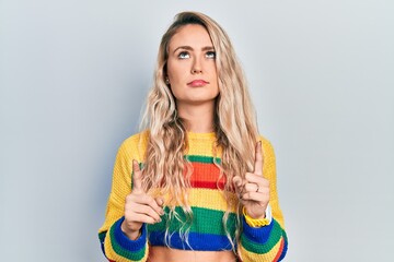 Beautiful young blonde woman wearing colored sweater pointing up looking sad and upset, indicating...