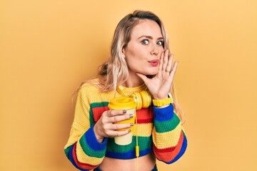 Beautiful young blonde woman drinking cup of coffee wearing headphones hand on mouth telling secret...