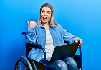 Beautiful caucasian woman sitting on wheelchair using laptop pointing thumb up to the side smiling happy with open mouth