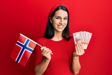 Young hispanic woman holding norway flag and krone banknotes smiling with a happy and cool smile on face. showing teeth.
