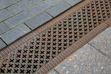 a drainage ditch covered with an iron patterned lattice on a sidewalk made of granite tiles stones...