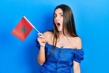 Young brunette teenager holding morocco flag scared and amazed with open mouth for surprise, disbelief face