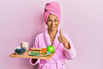 Middle age hispanic woman wearing bathrobe holding breakfast tray smiling happy and positive, thumb up doing excellent and approval sign