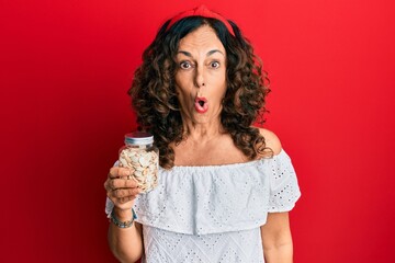 Middle age hispanic woman holding jar with pumpkin seeds scared and amazed with open mouth for surprise, disbelief face