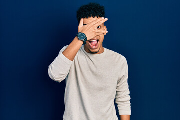 Fototapeta premium Young african american man wearing casual clothes peeking in shock covering face and eyes with hand, looking through fingers with embarrassed expression.
