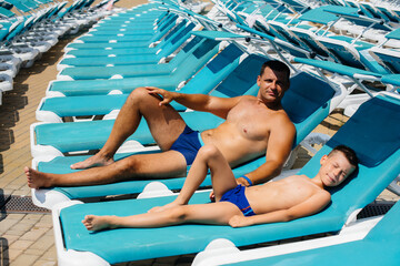 A young athletic man and his son are smiling happily and sunbathing on a sun lounger on a sunny...
