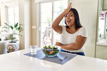 Young hispanic woman eating healthy salad at home surprised with hand on head for mistake, remember error. forgot, bad memory concept.