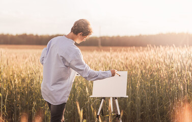 Cute young guy draws on canvas on an easel in nature in the summer. Artist on a glider