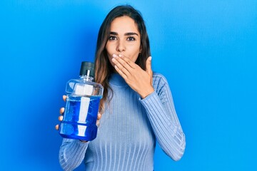 Young hispanic girl holding mouthwash for fresh breath covering mouth with hand, shocked and afraid...