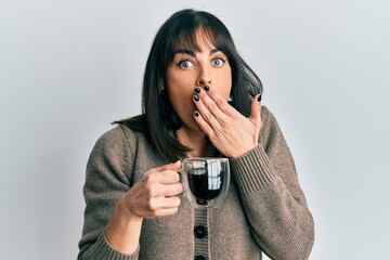 Young hispanic woman drinking a cup of coffee covering mouth with hand, shocked and afraid for...