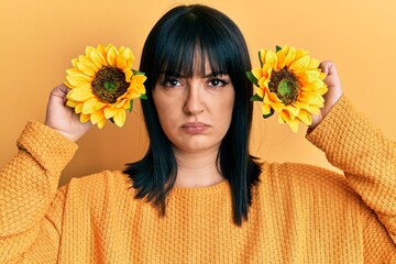 Young hispanic woman holding sunflowers over eyes skeptic and nervous, frowning upset because of...