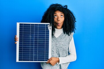 African american woman with afro hair holding photovoltaic solar panel making fish face with mouth...