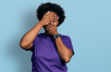 African american woman with afro hair wearing casual purple t shirt covering eyes and mouth with hands, surprised and shocked. hiding emotion