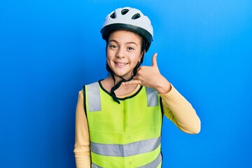 Beautiful brunette little girl wearing bike helmet and reflective vest smiling doing phone gesture with hand and fingers like talking on the telephone. communicating concepts.
