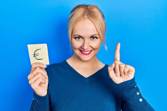 Young blonde woman holding euro symbol reminder smiling with an idea or question pointing finger with happy face, number one