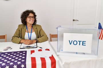 Young hispanic woman at political election sitting by ballot looking away to side with smile on...