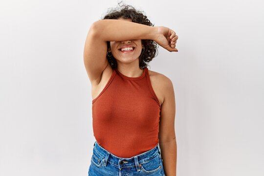 Young hispanic woman wearing glasses standing over isolated background covering eyes with arm smiling cheerful and funny. blind concept.