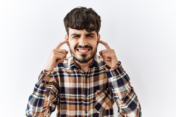 Hispanic man with beard standing over isolated background covering ears with fingers with annoyed expression for the noise of loud music. deaf concept.