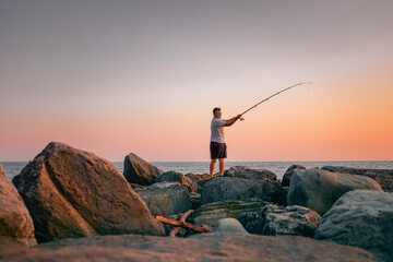 Obraz premium A man with a fishing rod is fishing on the rocks near the sea. Active recreation in nature. A fisherman catches fish at sunset.