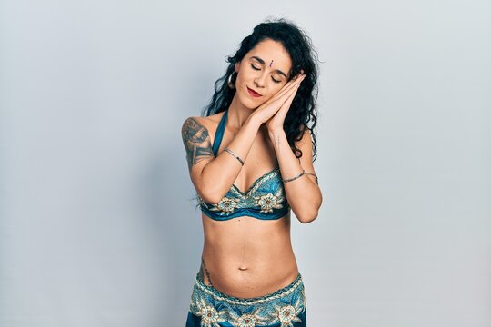 Young woman wearing bindi and traditional belly dance clothes sleeping tired dreaming and posing with hands together while smiling with closed eyes.