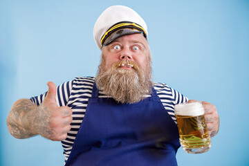 Joyful bearded person with overweight in sailor suit holds mug of tasty beer and shows thumb up on...