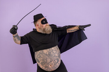Mature bearded fat man in Zorro costume with large bare abdomen stands in bellicose pose on purple...