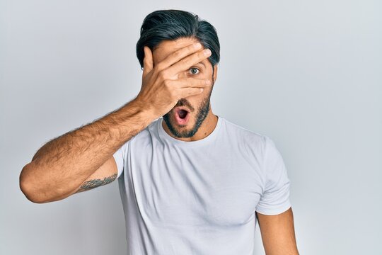 Young hispanic man wearing casual white t shirt peeking in shock covering face and eyes with hand, looking through fingers afraid