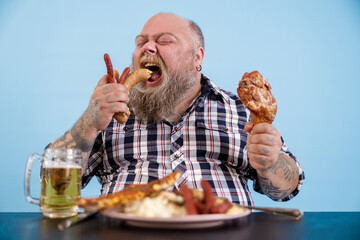 Hungry bearded plus size man eats sausages holding chicken leg at table with rich food and mug of beer on light blue background in studio