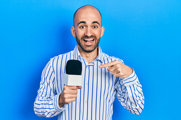 Young bald man holding reporter microphone smiling happy pointing with hand and finger