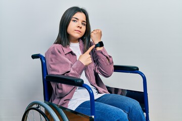 Young brunette woman sitting on wheelchair in hurry pointing to watch time, impatience, looking at the camera with relaxed expression