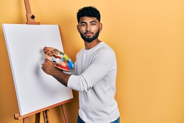 Arab man with beard standing drawing with palette by painter easel stand relaxed with serious expression on face. simple and natural looking at the camera.