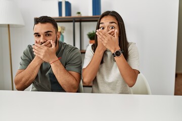 Young latin couple wearing casual clothes sitting on the table shocked covering mouth with hands for mistake. secret concept.