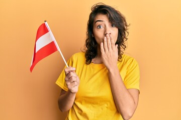 Young hispanic woman holding austria flag covering mouth with hand, shocked and afraid for mistake....