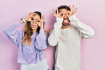 Young hispanic couple wearing casual clothes doing ok gesture like binoculars sticking tongue out,...