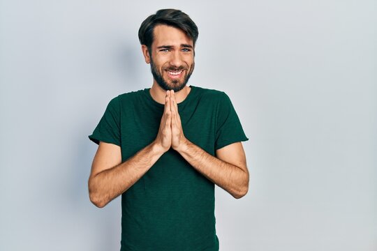 Young hispanic man wearing casual white tshirt praying with hands together asking for forgiveness smiling confident.