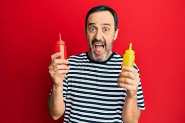 Middle age hispanic man holding ketchup and mustard bottle celebrating crazy and amazed for success with open eyes screaming excited.