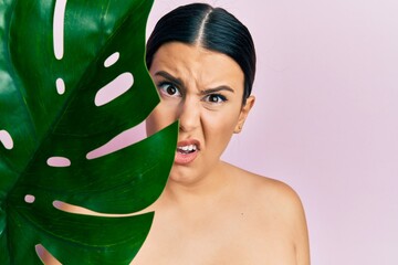 Beautiful brunette woman holding green plant leaf close to beautiful face clueless and confused...