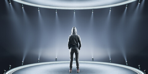 Back view of young hacker in hoodie standing on creative stage background with illumination....