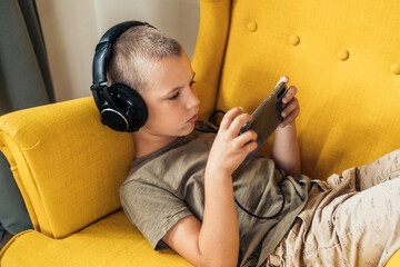 Young boy lying in the armchair and watching video in smartphone in headphones. Internet communication, modern technologies and people concept.
