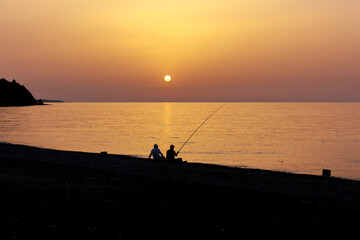 Silhouette of two men fishing on the beach during the sunrise. Sun rises over the Mediterranean Sea on a lovely summer morning. Selective focus.
