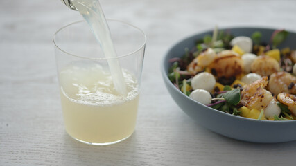 Man pour lemonade into tumbler glass next to salad with shrimps, mango, mozzarella and micro greens