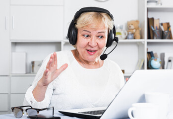 Naklejka premium Buxom woman of an age sits in front of table with laptop in headphones with microphone.