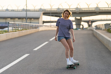Active woman longboarder of middle age riding longboard on empty road or highway. Casual urban female relaxing on skateboard after work. Lady of 40s skateboarding at sunset. Modern lifestyle concept