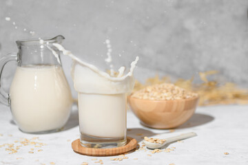 Splash of oat milk in a glass and jug on a grey stone table background and a bowl with oat flake. Jug with vegan non dairy alternative milk.