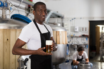 African american man brewer in apron standing with glass of beer in brew-house, man on backround