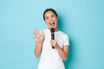 Lifestyle, people and leisure concept. Cheerful smiling asian girl enjoying karaoke night. Japanese female student singing song in microphone with happy emotions, standing light blue background - obrazy, fototapety, plakaty