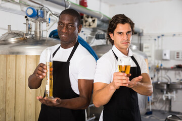 Two professional brewers showing glass and flask of beer in brewer factory