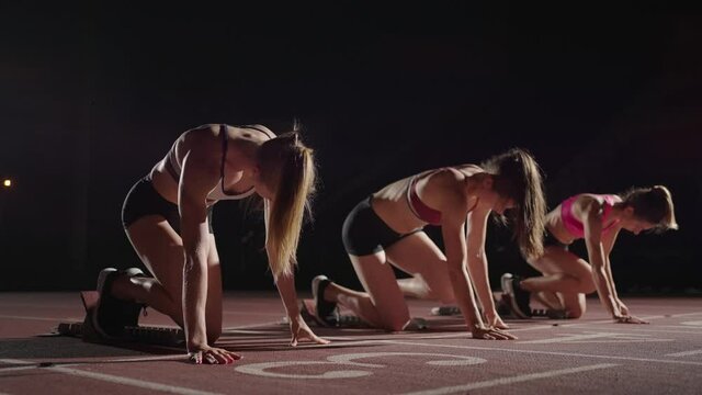 Slow motion 3 female runners running to the camera in a sports arena. Track and field athletics competition running in the dark. Race together