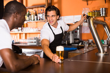 Positive man bartender giving glass of golden beer to man client in bar
