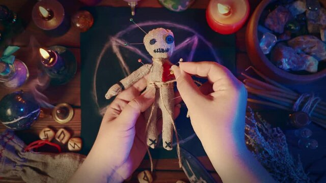 Sorceress or witch sticks needles into voodoo doll at ritual table with pentagram, burning candles and other occult objects, top view. Voodoo witchcraft, spirituality and occultism concept.